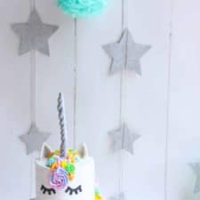 A white cake decorate to look like a unicorn on a white backdrop with sparkly stars hanging in the back