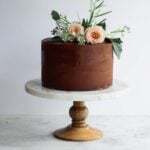 A chocolate cake on a marble and wood cake stand with flower decorations
