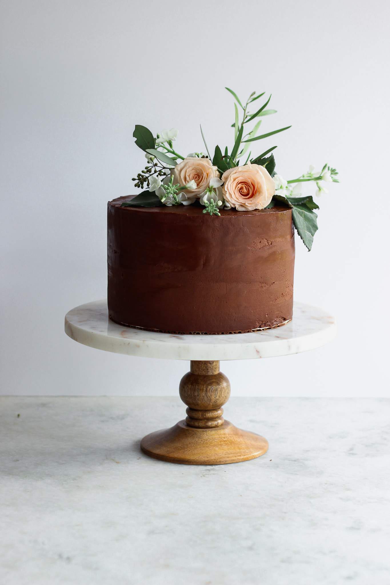 A chocolate cake on a marble and wood cake stand with flower decorations