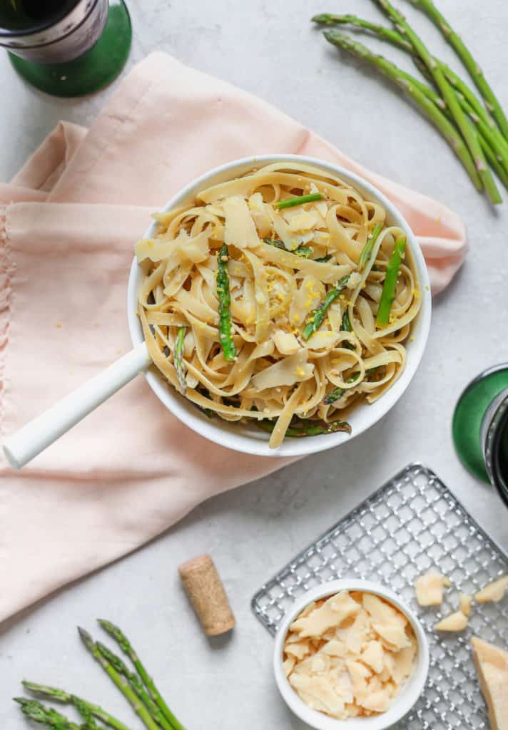 Fettuccine pasta with asparagus and lemon served in a white bowl over a pink napkin with parmesan cheese on the side.