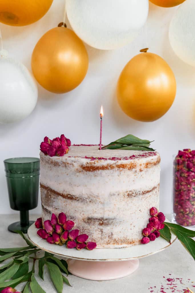 orange cardamom cake garnished with dried rose petals and lit candle on cake stand in front of balloon backdrop
