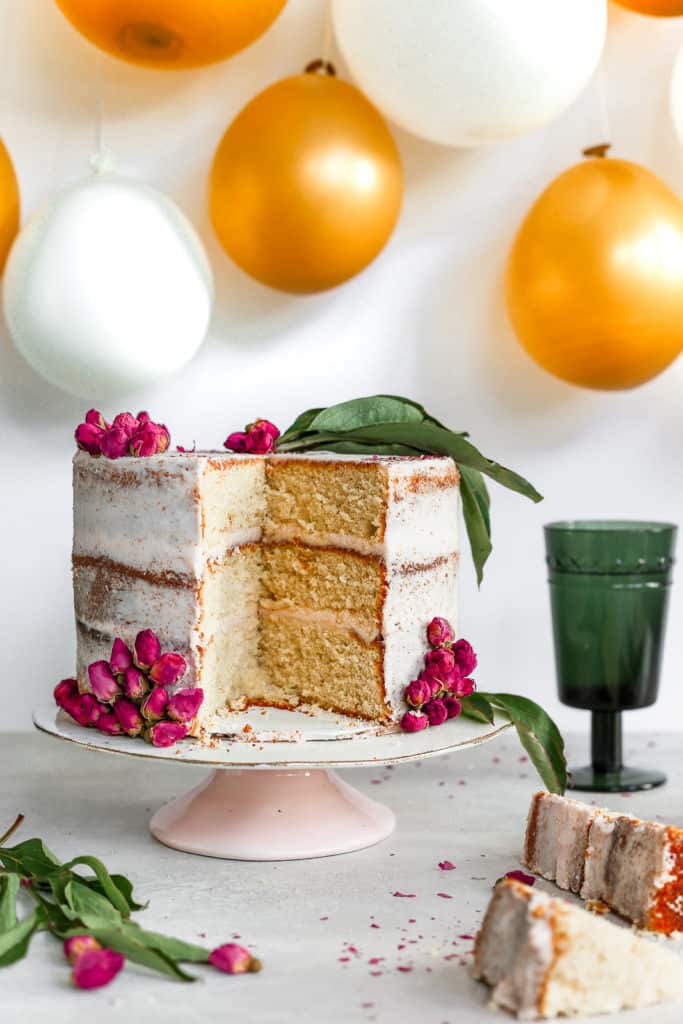 sliced orange cardamom cake garnished with dried rose petals on cake stand in front of balloon backdrop