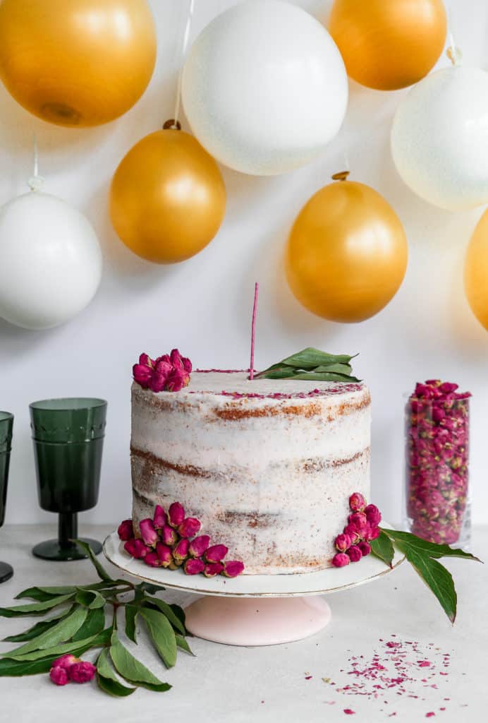 orange cardamom cake garnished with dried rose petals on cake stand in front of balloon backdrop