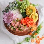 A white cake on a brown cake stand decorated with a rainbow of fresh flowers