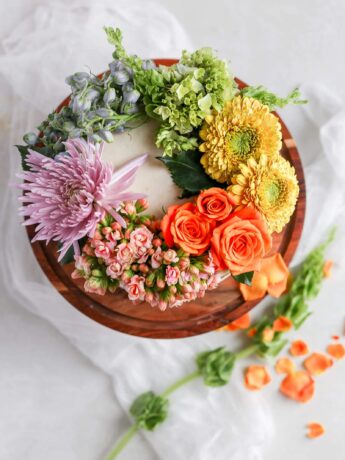 A white cake on a brown cake stand decorated with a rainbow of fresh flowers