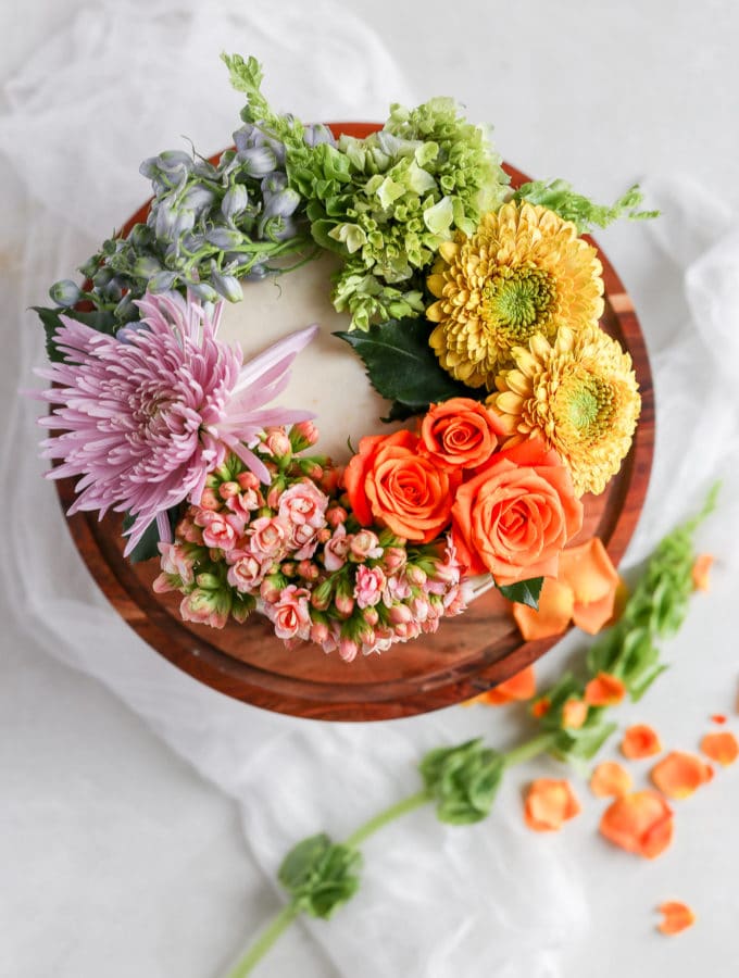 A white cake on a brown cake stand decorated with a rainbow of fresh flowers
