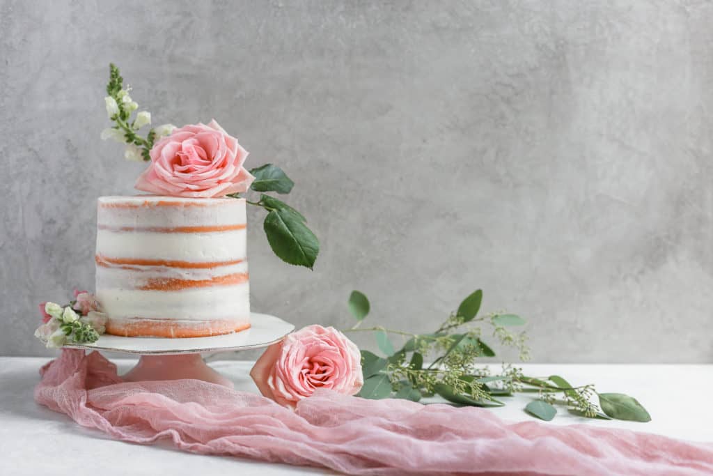 A naked cake decorated with pink roses on a cake stand with pink cheesecloth on a gray board