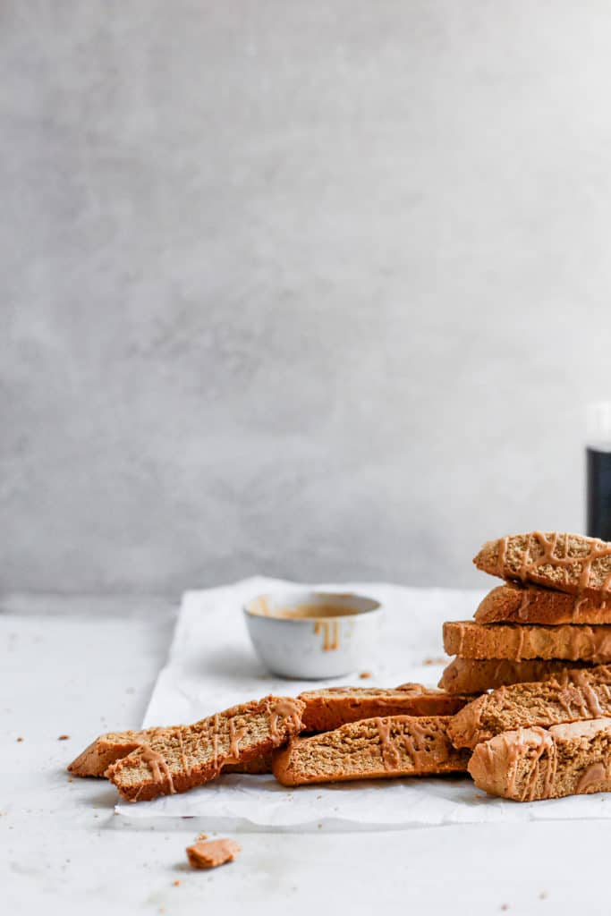 Cookie butter biscotti layered on top of each other on parchment paper with a bowl of glaze in the background.