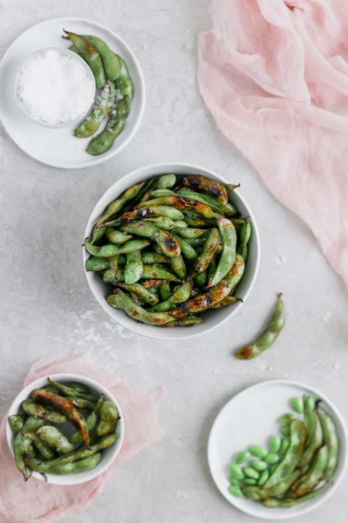 Blistered edamame with soy sauce and lime in a bowl styled with pink cheesecloth on a gray background.