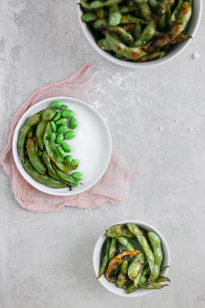 Blistered edamame with soy sauce and lime in a bowl styled with pink cheesecloth on a gray background.