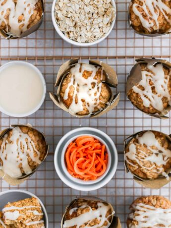 Carrot cake muffins with cream cheese glaze styled with bowls of ingredients on a cooling rack.
