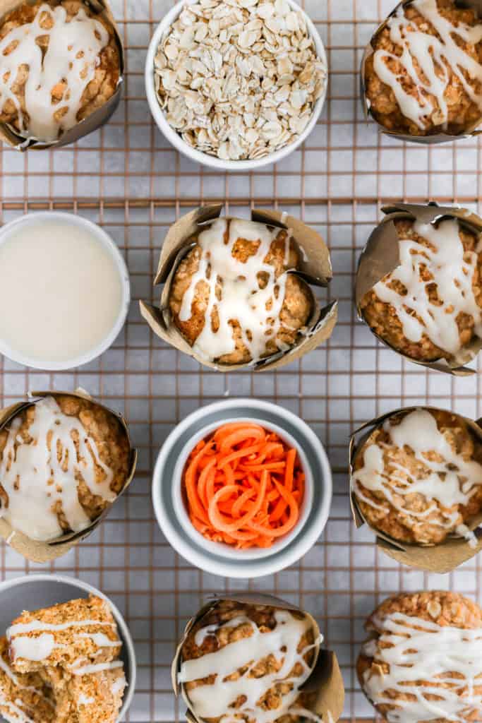 Carrot cake muffins with cream cheese glaze styled with bowls of ingredients on a cooling rack.