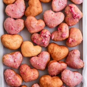 A mixture of strawberry glazed and plain fried heart shaped donuts on a sheet pan.