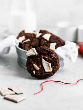 A chocolate peppermint cookie leaning against a tin of other cookies next to red string.