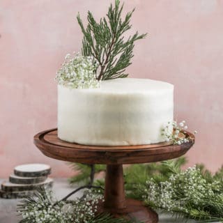 Cream cheese frosted red velvet cake decorated with leaves on a wood cake stand on a pink background.