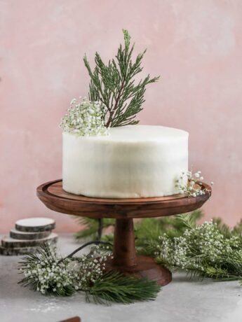 Cream cheese frosted red velvet cake decorated with leaves on a wood cake stand on a pink background.