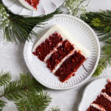 A slice of red velvet cake on a small gray plate with a gold fork styled on a gray background.