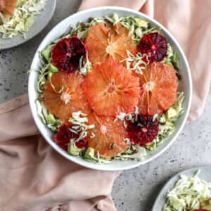 A citrus cabbage salad with grapefruit, blood oranges, cabbage, avocado, and sunflower seeds in a white bowl styled on a pink napkin on a gray surface.