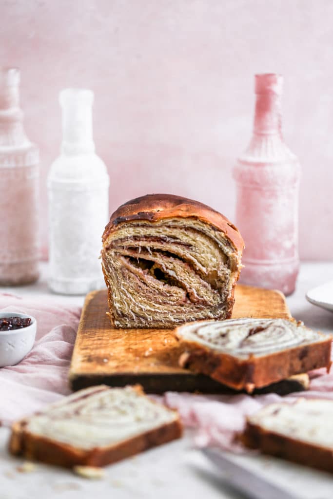 Cheese babka with a raspberry jam swirl sliced sitting on tiop of a cutting board on a pink background. 