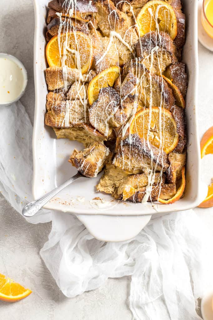 A spoon dipping into a baked casserole with challah and oranges and drizzled with white chocolate