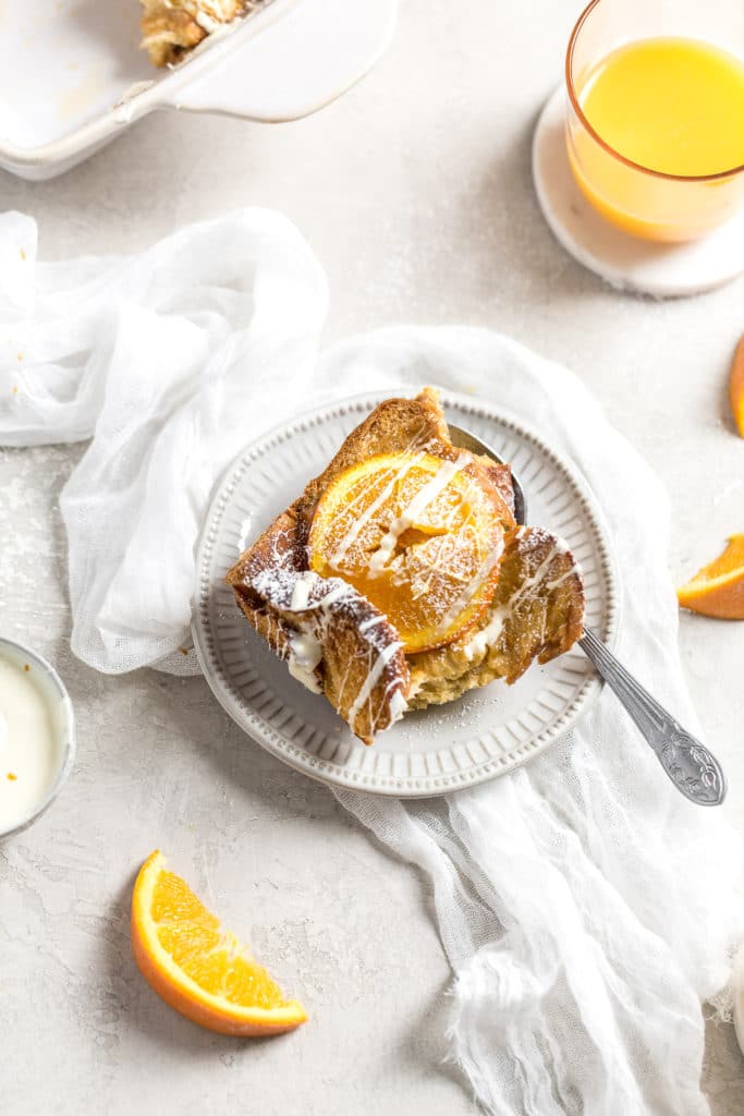 A serving of challah French toast with and orange on top on a plate with orange juice in a cup in the background