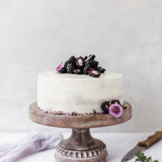 White cake on a brown cake stand with purple flowers and blackberries