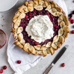 Cranberry pie with whipped cream resting on a white linen on a gray backdrop with a knife lying on an angle.