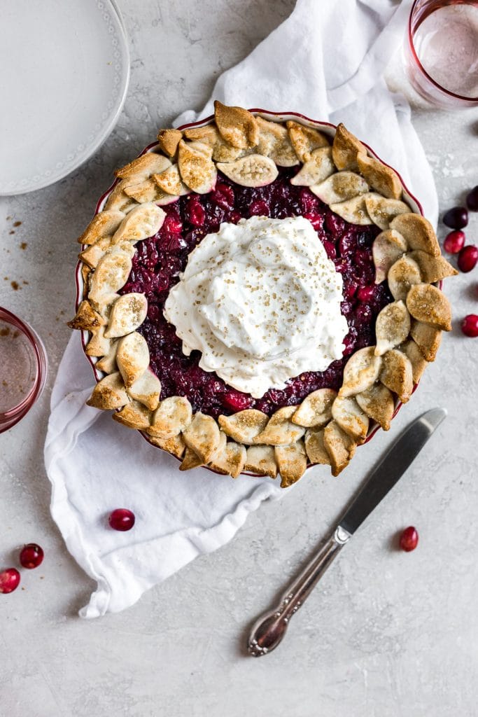 Cranberry pie with whipped cream resting on a white linen on a gray backdrop with a knife lying on an angle.