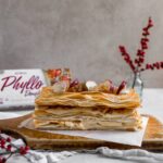 A Napoleon dessert made with phyllo dough on a wooden cutting board topped with dried apples and red flower berries in the background.