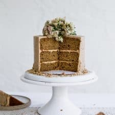 A brown cake with beige frosting that has a few slices taken out of it sitting on a white cake stand on a white background
