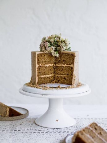 A brown cake with beige frosting that has a few slices taken out of it sitting on a white cake stand on a white background