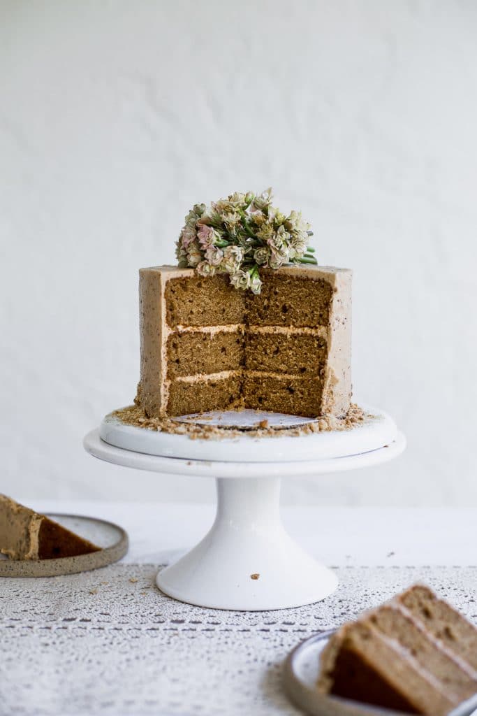 A brown cake with beige frosting that has a few slices taken out of it sitting on a white cake stand on a white background 