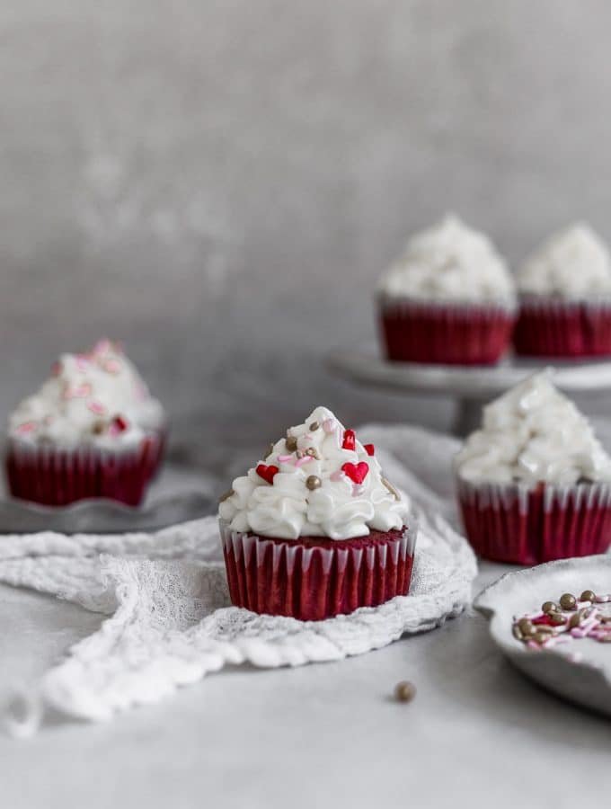 a red velvet cupcake with cream cheese frosting sitting on white gauze with other cupcakes in the background on a gray surface.