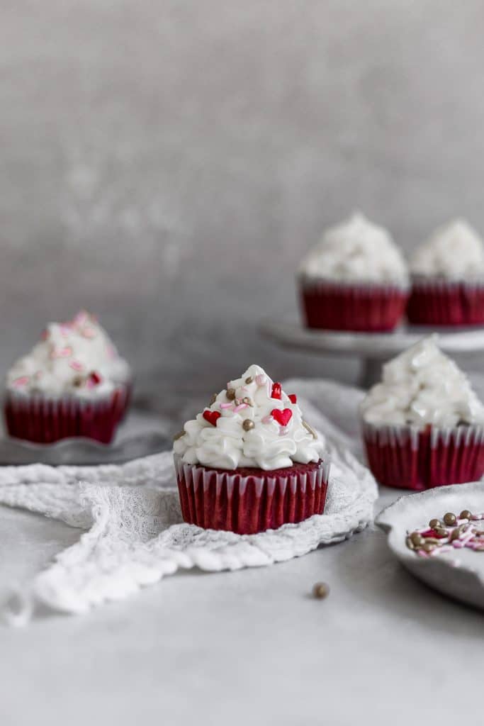 a red velvet cupcake with cream cheese frosting sitting on white gauze with other cupcakes in the background on a gray surface.