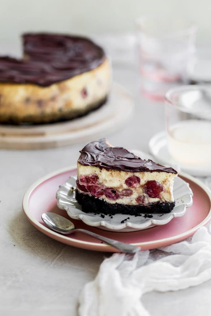 A slice of chocolate cheesecake with cherries in the middle sitting on two plates. The larger one is pink the smaller one is gray. There is a spoon on the pink plate. 