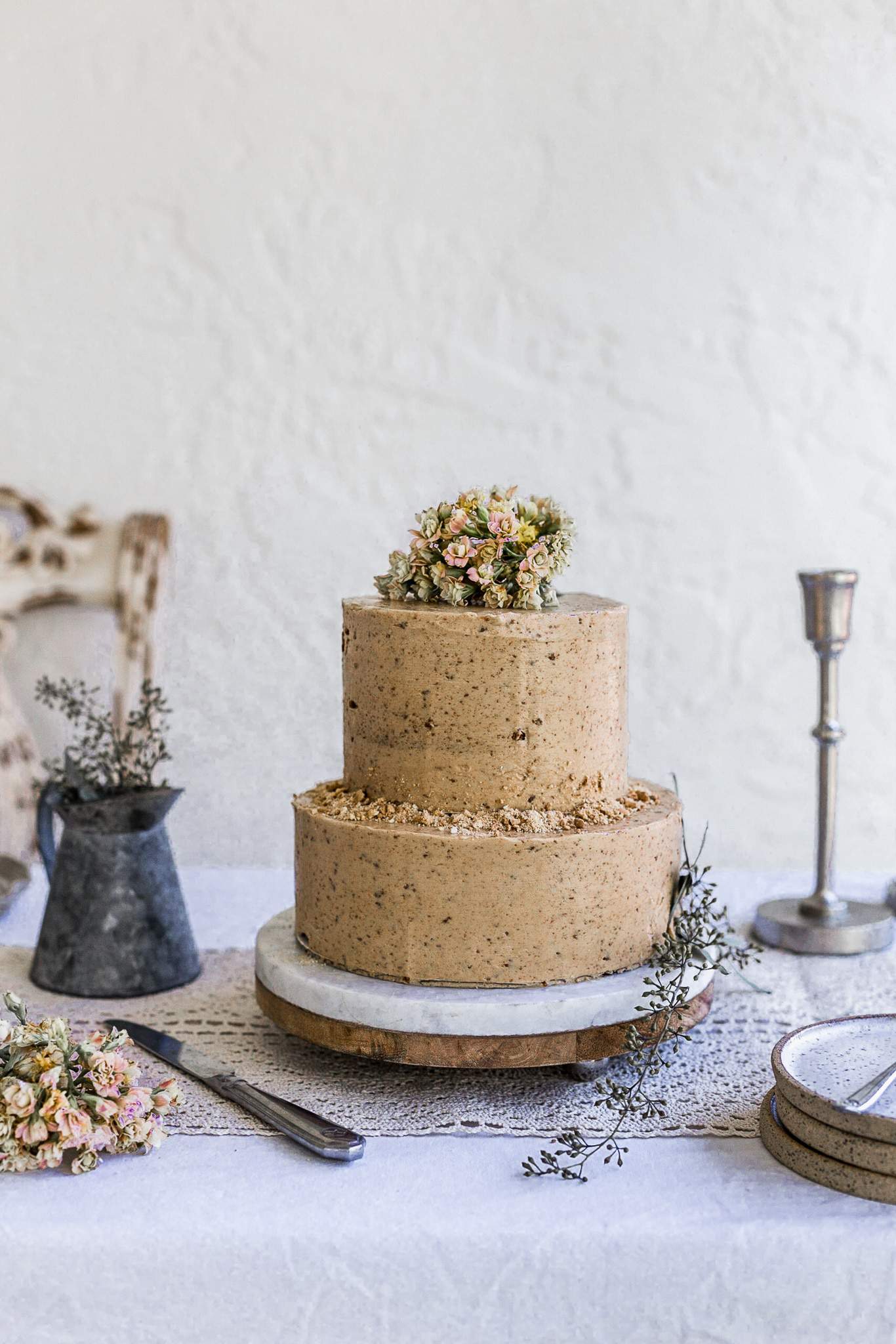 A two tier cookie butter cake on a marble and wood cake plate sitting on a white eyelet tablecloth decorated with pink flowers on top.