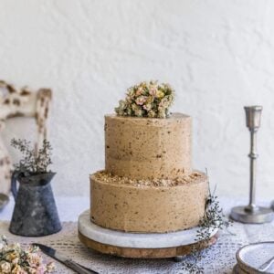 A two tier cake with brown frosting sitting on a decorated table with flowers on top