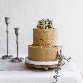 A two tier cake with light brown frosting sitting on a marble cake stand on a white table with candlesticks in the background