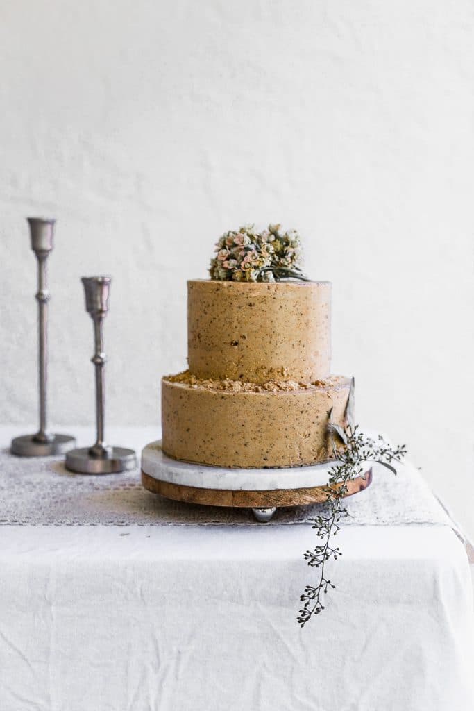 A two tier cake with light brown frosting sitting on a marble cake stand on a white table with candlesticks in the background