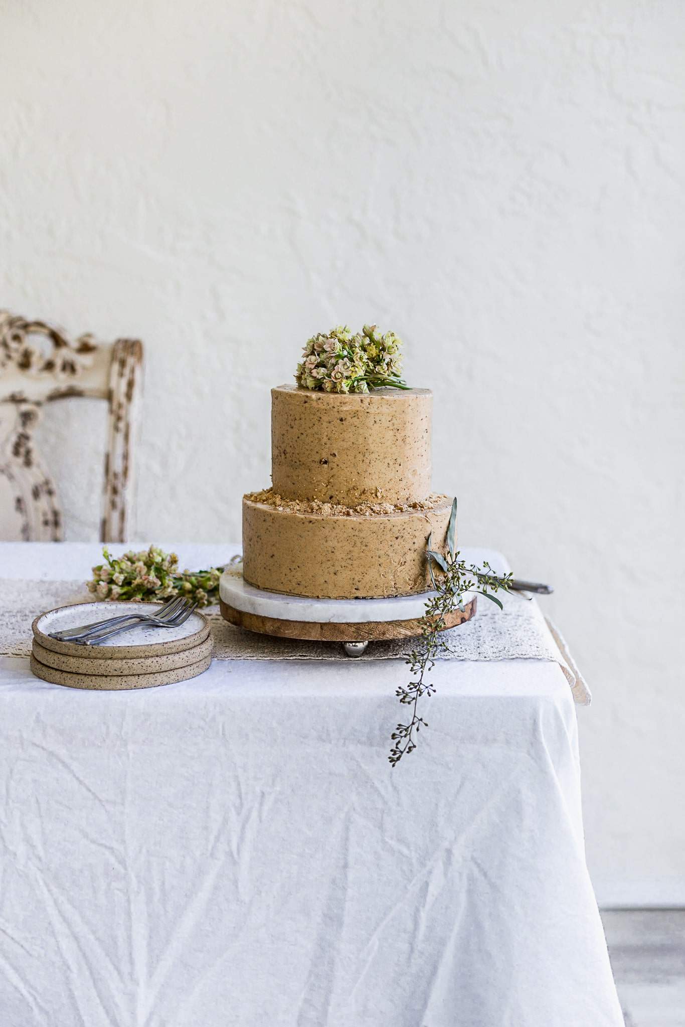 A two tier brown cake decorated with flowers and cookie crumbs sitting on a white table with plates and forks on the side.