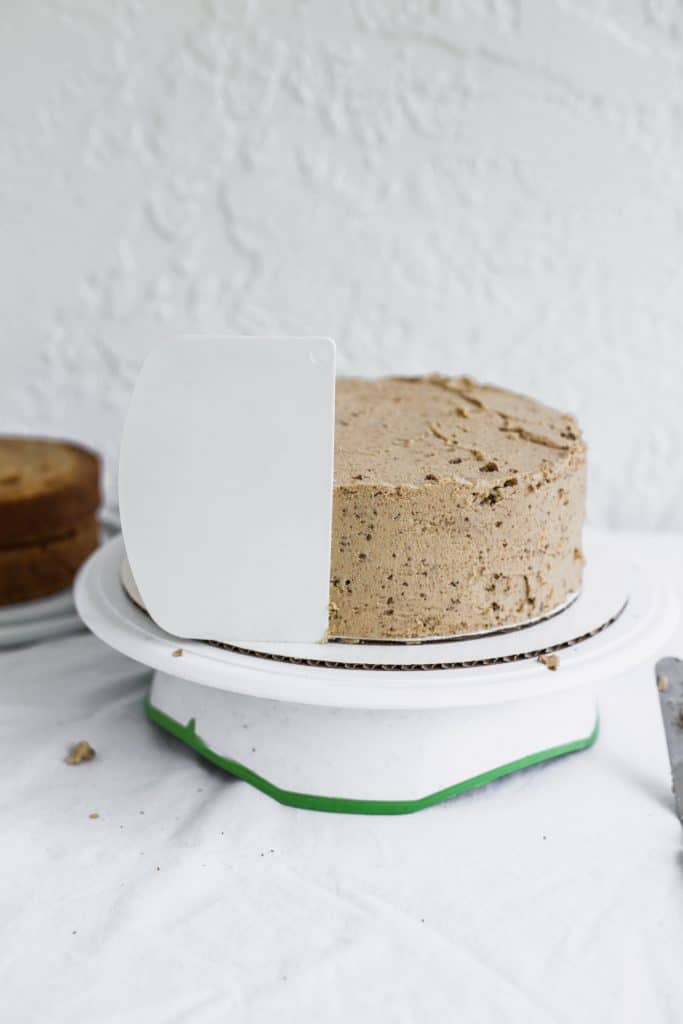 A white bench scraper sticking to the side of a beige frosted cake 