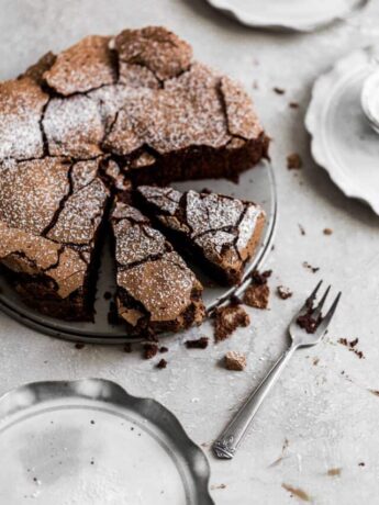 The top of a chocolate cake with 2 slices cut out with cracked top and a dusting of confectioners sugar on top on a gray background with a fork next to it.