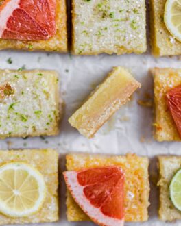 A lemon bard on its side showing the texture next to other cut squares of lemon bars topped with slices of grapefruit and lemons
