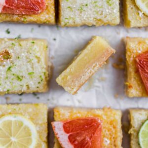 A lemon bard on its side showing the texture next to other cut squares of lemon bars topped with slices of grapefruit and lemons