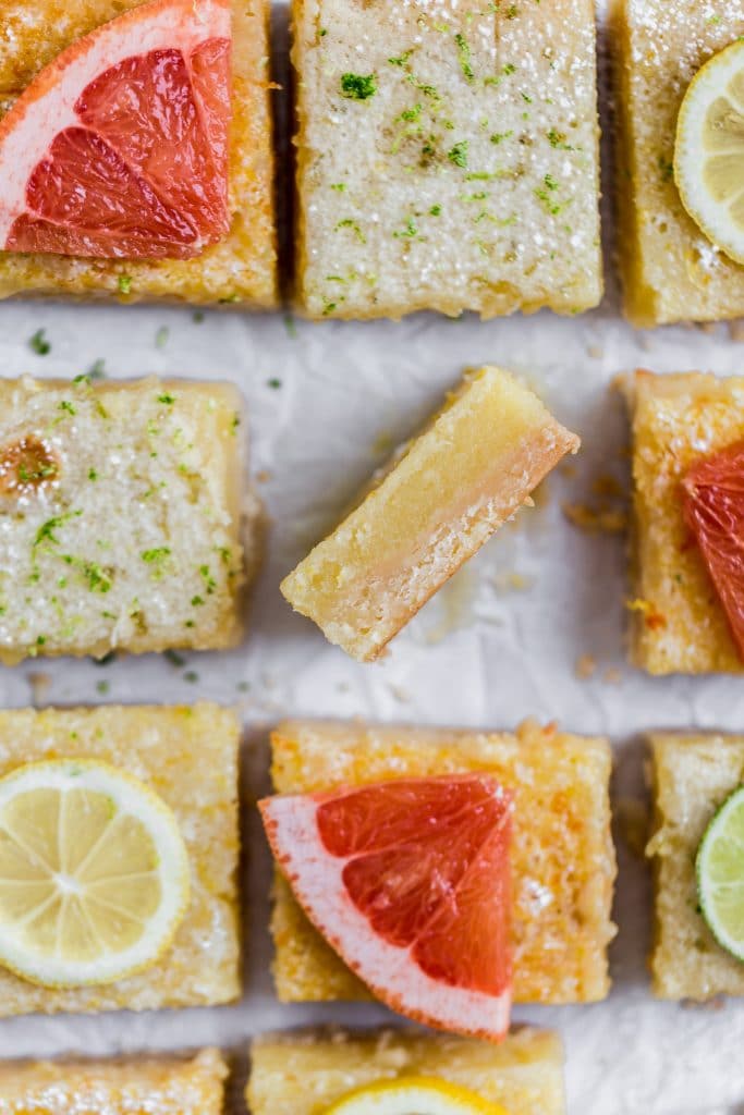 A lemon bard on its side showing the texture next to other cut squares of lemon bars topped with slices of grapefruit and lemons