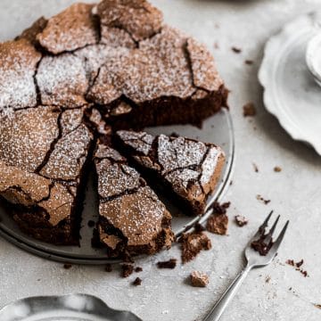 The top of a chocolate cake with 2 slices cut out with cracked top and a dusting of confectioners sugar on top on a gray background with a fork next to it.