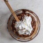 Whipped egg whited on top of chocolate batter in a glass bowl with a spatula sticking out
