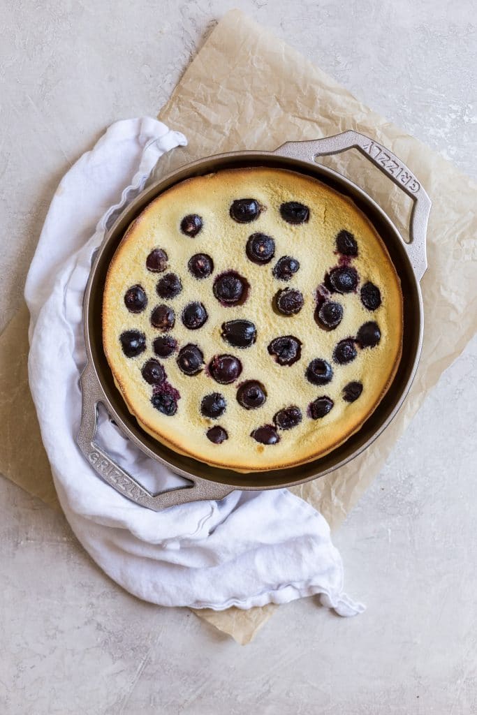 A yellow clafoutis recipe with cherries in it baked in a cast iron pan on a gray surface