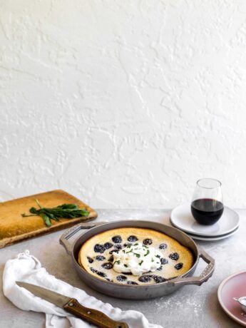 The side view of a cherry clafoutis made in a GRIZZLY nickel plated cast iron pan set on a gray table with a cutting board in the background and a cup of wine on some plates.