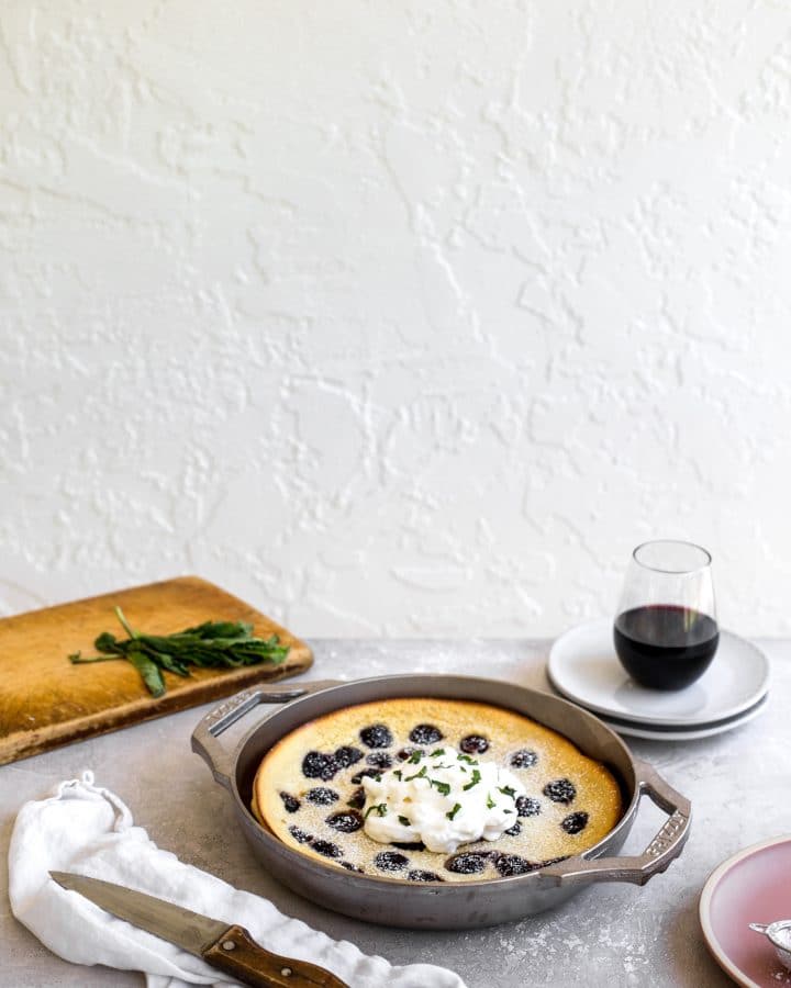 The side view of a cherry clafoutis made in a GRIZZLY nickel plated cast iron pan set on a gray table with a cutting board in the background and a cup of wine on some plates.